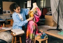 Estilista em uma oficina de moda criando um vestido colorido, com tecidos, moldes, acessórios e manequim ao redor