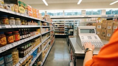 Visão em primeira pessoa de um supermercado organizado, com prateleiras abastecidas, caixa registradora e um jogador administrando a loja.